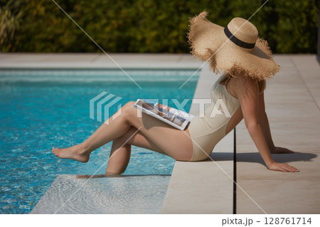 Woman in stylish swimsuit relaxing by poolside with magazine and sun hat enjoying summer vibes 128761714