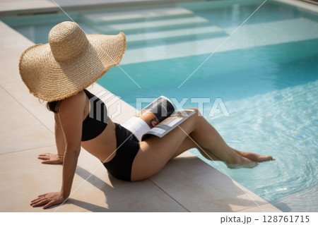 Woman relaxing by poolside, wearing large sunhat, reading magazine, enjoying summer leisure time 128761715