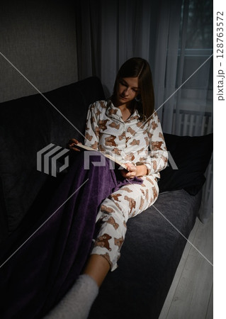 Young woman reading book in pajamas on sofa at night. Young woman reading book in pajamas on sofa at night. 128763572