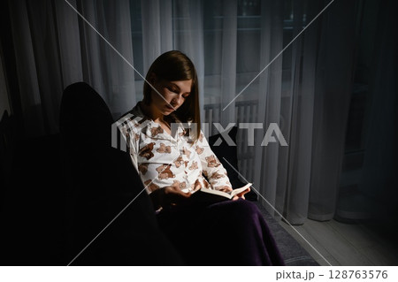 Young woman reading book in pajamas on sofa at night. 128763576