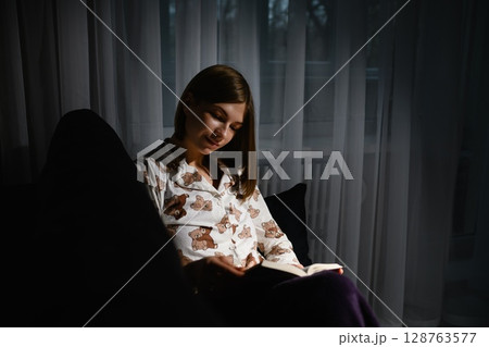 Young woman reading book in pajamas on sofa at night. 128763577