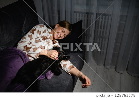 Woman cuddling black cocker spaniel puppy on sofa at night. 128763579