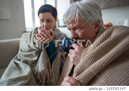 Sad elderly couple under blanket drinking hot tea, suffer from illness at home. Infection quarantine Sad elderly couple under blanket drinking hot tea, suffer from illness at home. Infection quarantine 128765147