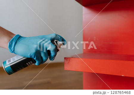 Housewife wearing blue gloves applying scotch remover spray to dried adhesive stain on red shelf, performing thorough housekeeping at home, close-up. Concept of easy housekeeping domestic life. 128765468