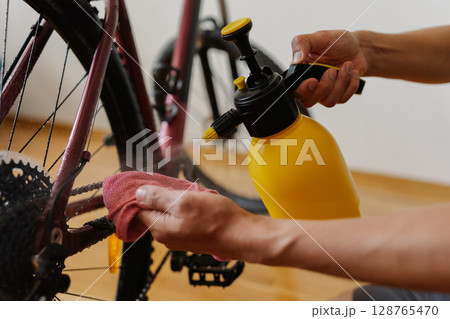 Detail cropped shot of unrecognizable man using yellow sprayer and cloth preparing to clean chain of mountain bike, emphasizing importance of bicycle maintenance after trail riding at home. Detail cropped shot of unrecognizable man using yellow sprayer and cloth preparing to clean chain of mountain bike, emphasizing importance of bicycle maintenance after trail riding at home. 128765470