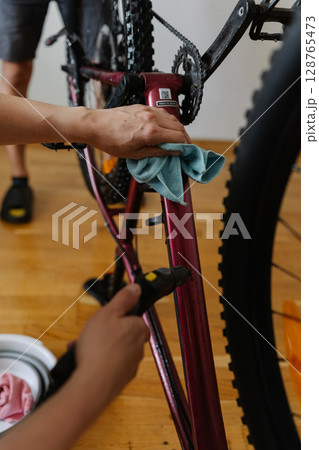 Two people cleaning mountain bike frame using water sprayer and cloth, removing dirt and grime performing detailed maintenance after trail riding, preserving cycling gear condition and functionality. Two people cleaning mountain bike frame using water sprayer and cloth, removing dirt and grime performing detailed maintenance after trail riding, preserving cycling gear condition and functionality. 128765473