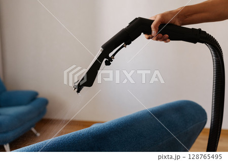 Detail cropped shot of unrecognizable homemaker removing dirt and stains using washing vacuum cleaner to clean blue sofa in living room, demonstrating modern approach to household chores. 128765495