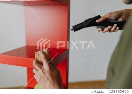 Close-up of professional janitor male cleaning red shelf using modern steam cleaner and sponge, ensuring hygiene and order in commercial or residential setting. Concept of housekeeping domestic life. Close-up of professional janitor male cleaning red shelf using modern steam cleaner and sponge, ensuring hygiene and order in commercial or residential setting. Concept of housekeeping domestic life. 128765496