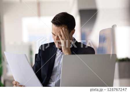 Businessman reading document with shocked or distressed expression at workplace 128767247