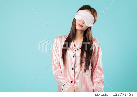 Asian woman in pajamas, portrait with eye mask cover for sleep, young beauty follows her routine to rest and relax, planning to take Sunday nap hoping for good sleep, isolated studio blue background 128772455