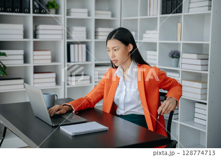 Beautiful Asian woman using laptop and tablet while sitting at her working place. Concentrated at work. Beautiful Asian woman using laptop and tablet while sitting at her working place. Concentrated at work. 128774713