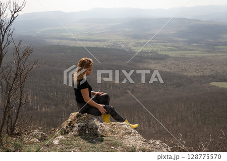 Woman Hillside Landscape: Blonde woman sits on a rock, viewing the vast valley landscape, daytime. 128775570