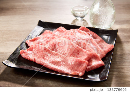 黒毛和牛の霜降りスライス肉を黒い角皿に美しく盛り付け、日本酒の徳利と盃を添えた高級感あふれる和食 128776693