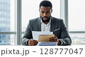 A concerned businessman reviews important documents, his expression reflecting worry and surprise at his desk near a large window. 128777047