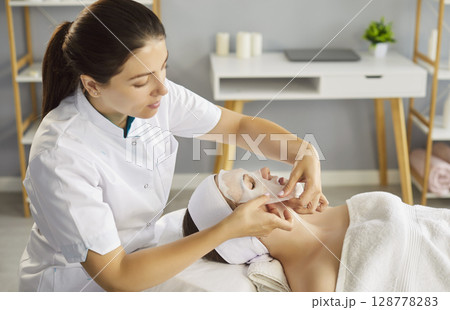 Young attractive cosmetologist woman applying sheet mask on female face in salon Young attractive cosmetologist woman applying sheet mask on female face in salon 128778283
