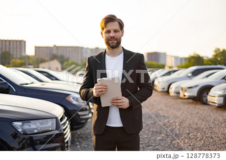 A car salesman in a suit stands in a car parking with a tablet. 128778373