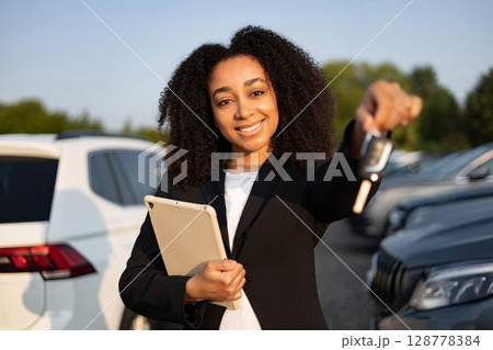 A smiling woman in a car dealership is selling a car, holding the keys and a tablet. 128778384