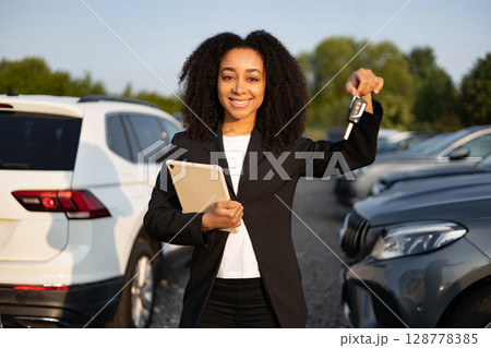 An African American woman, car salesperson, smiles while holding car keys and a tablet with vehicles visible in the background. An African American woman, car salesperson, smiles while holding car keys and a tablet with vehicles visible in the background. 128778385