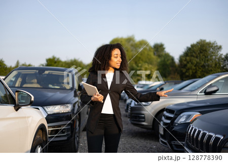 A woman, presumably a car salesperson, points to a vehicle in a car lot while holding a tablet, presenting various car models for sale. A woman, presumably a car salesperson, points to a vehicle in a car lot while holding a tablet, presenting various car models for sale. 128778390