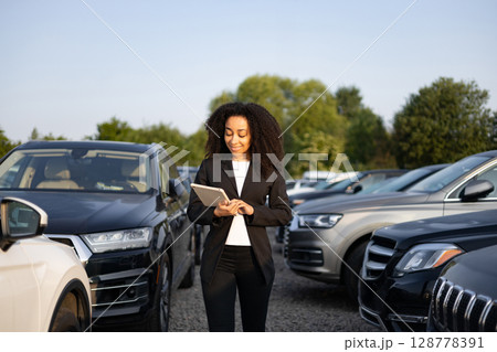 A woman in a suit is smiling while using a tablet in a car dealership. A woman in a suit is smiling while using a tablet in a car dealership. 128778391