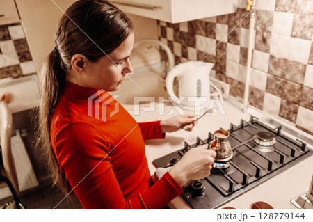 Woman Kitchen Coffee Brewing: Home morning routine, young woman preparing Turkish coffee on gas stove. Woman Kitchen Coffee Brewing: Home morning routine, young woman preparing Turkish coffee on gas stove. 128779144