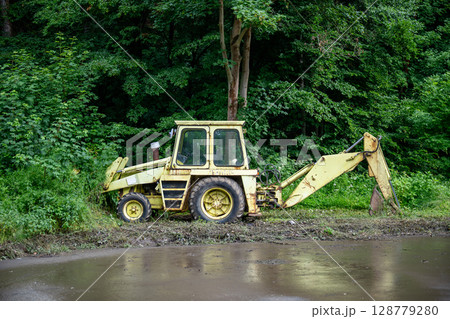 Old yellow backhoe loader parked at forest edge with overgrown vegetation Old yellow backhoe loader parked at forest edge with overgrown vegetation 128779280