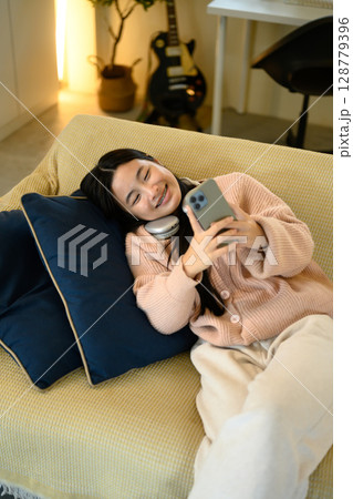 Teenage girl lying on the couch, scrolling through social media on her phone 128779396