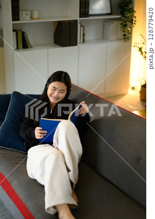 Teenage girl sitting on a sofa, reading a book and relaxing at home 128779420