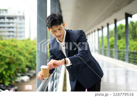 Businessman with takeaway coffee checking time on wrist watch during busy workday Businessman with takeaway coffee checking time on wrist watch during busy workday 128779518