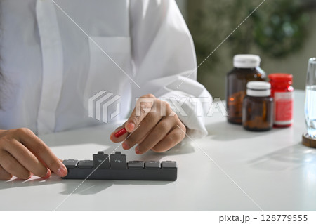 Close up of woman organizing daily medication into a pill box on a white desk 128779555