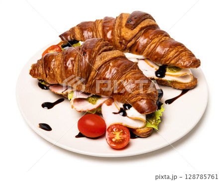 Two freshly baked classic delicious fresh organic croissants with ham, cheese, pesto sauce and tomatoes on a white ceramic plate isolated on white background Two freshly baked classic delicious fresh organic croissants with ham, cheese, pesto sauce and tomatoes on a white ceramic plate isolated on white background 128785761