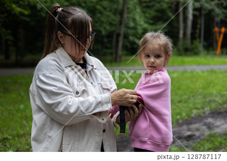 Mother cares about the child, putting on the protection of her daughter, preparing to skating on a bicycle in a bright green park, creating a mental moment of family connection. Maternal care. Mother cares about the child, putting on the protection of her daughter, preparing to skating on a bicycle in a bright green park, creating a mental moment of family connection. Maternal care. 128787117