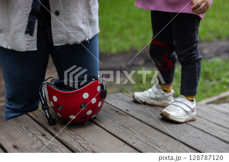 The red helmet lies on wooden boards, near the legs of the child, dressed in complex protection. The child is going to engage in a bicycle or scooter in the fresh air, emphasizing safety and fun. 128787120