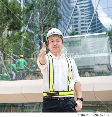 With a bright smile and a thumbs up a smiling Asian construction professional acknowledges success at a contemporary building site. With a bright smile and a thumbs up a smiling Asian construction professional acknowledges success at a contemporary building site. 128787485