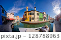 Brightly colored houses reflect in the serene canal of Burano, Venice during a clear sunny day 128788963
