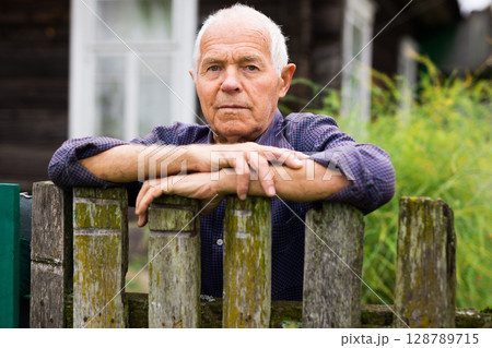 Portrait of pensive elderly man at fence of his country house in village Portrait of pensive elderly man at fence of his country house in village 128789715