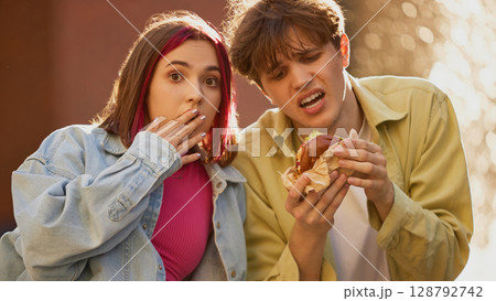 Shocked girl and disgusted boy reacting to strange burger outdoors Shocked girl and disgusted boy reacting to strange burger outdoors 128792742