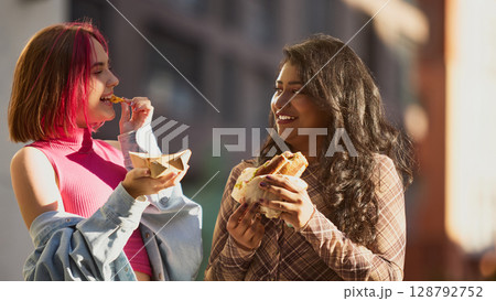 Two girls smiling and sharing fries and sandwich in bright daylight Two girls smiling and sharing fries and sandwich in bright daylight 128792752