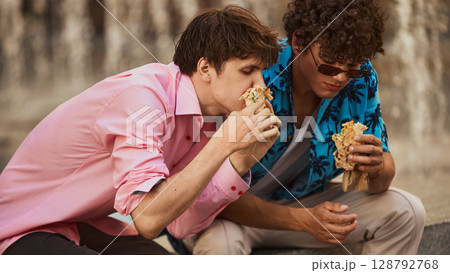 Two young men eating shawarma on city steps in casual vibrant clothing 128792768