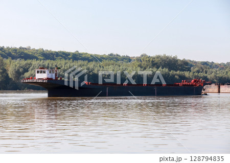 A large cargo ferry navigating calm waters of a river with a forested background 128794835