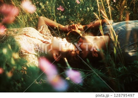 A couple lies together in a vibrant wildflower field, surrounded by colorful blooms and soft grass. They share a serene moment as sunlight casts a warm glow on their faces 128795497