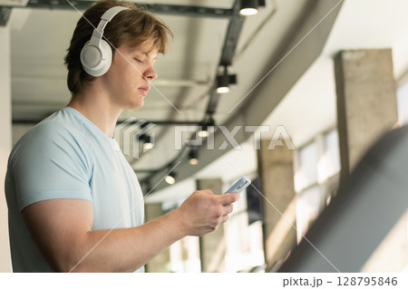 Athlete man with cellphone or mobile phone running track on treadmill at gym. Warming up. Preparation for cardio training. Work out at fitness center. Close up. Athlete man with cellphone or mobile phone running track on treadmill at gym. Warming up. Preparation for cardio training. Work out at fitness center. Close up. 128795846