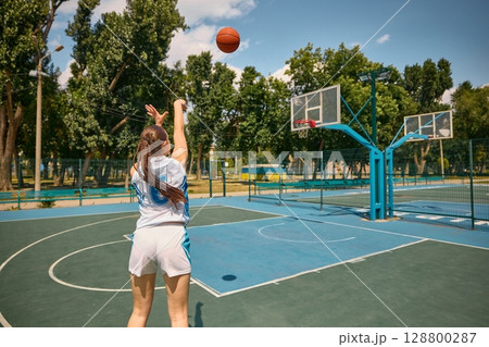 Young girl playing basketball on outdoor court beneath blue sky 128800287