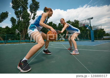 Female basketball athletes training dribbling skills on open-air court beneath cloudless sky Female basketball athletes training dribbling skills on open-air court beneath cloudless sky 128800317