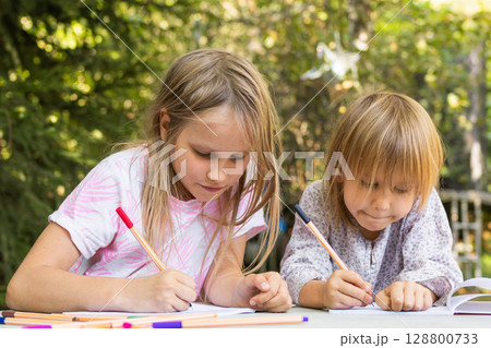 Two young sisters drawing with colorful fineliners in notebooks at an outdoor table Two young sisters drawing with colorful fineliners in notebooks at an outdoor table 128800733