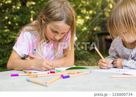 Two sisters drawing with fineliners in notebooks at a table in a summer garden Two sisters drawing with fineliners in notebooks at a table in a summer garden 128800734