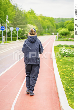Woman in black sportswear walks alone on red path in lush spring park. Concept of light running, healthy life, recreation, wellness walk, sport outdoor training 128800950