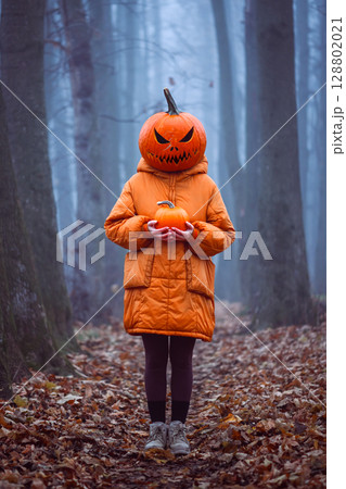 Halloween girl in carved pumpkin mask stands in foggy fall forest 128802021