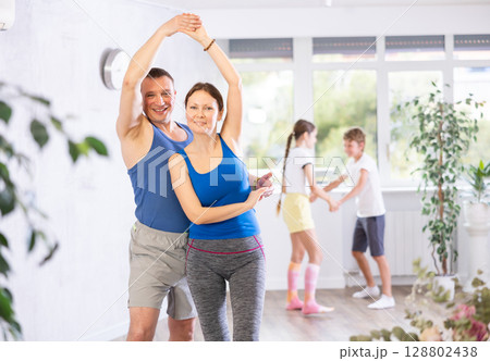 Cheerful friendly man and woman practicing active dance in pairs in front of their kids during family dance class in hall indoors 128802438