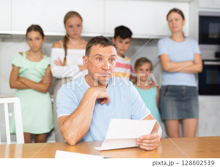 Frustrated dad is sitting in kitchen, reading letter. Blurred worried family in background 128802539
